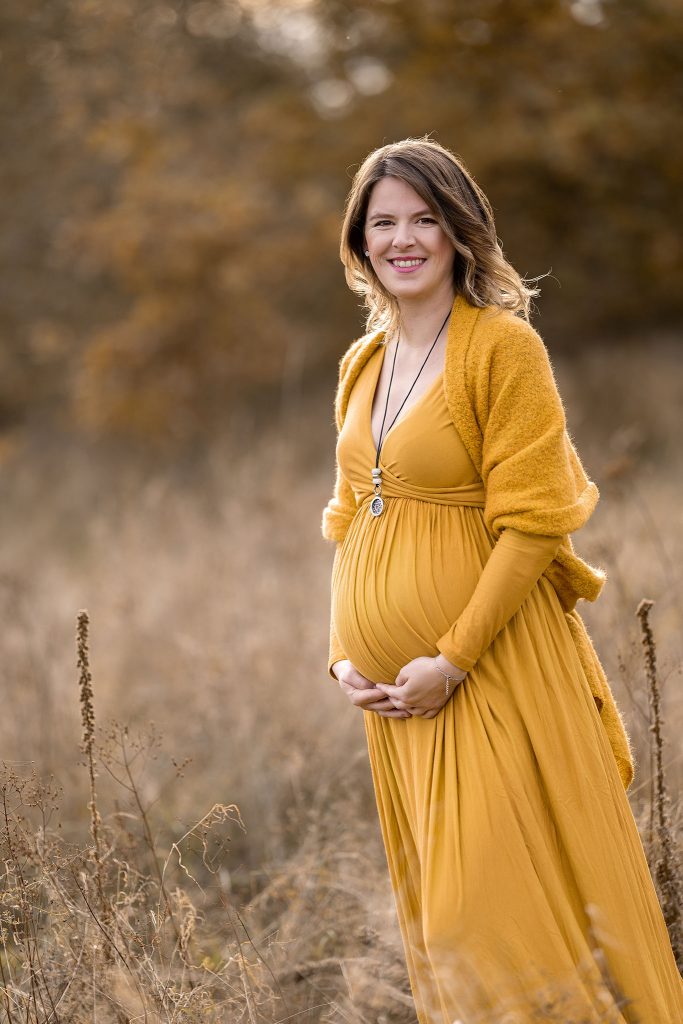 Schwangere in gelbem Kleid in einer Herbstlandschaft. Sie hält den Bauch mit beiden Händen und strahlt in die Kamera