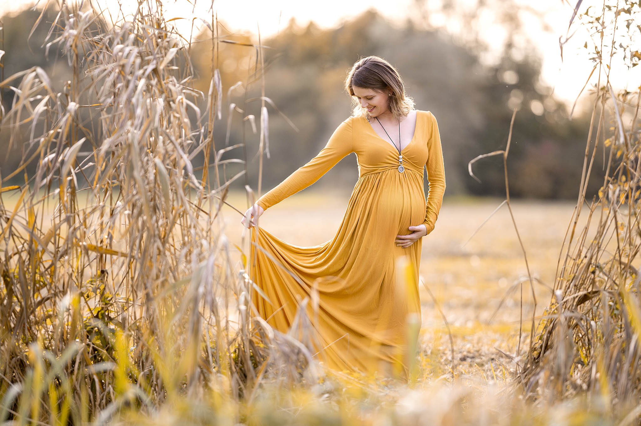 Schwangere in gelbem Kleid in einer Herbstlandschaft. Sie zieht das Kleid in Position und lächelt dabei.
