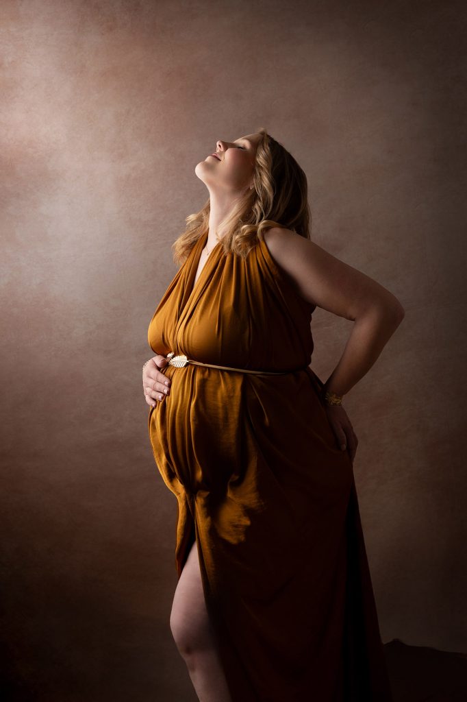 Schwangere Frau in goldenem griechischem Kleid, Blick anmutig nach oben gerichtet, Ausdruck von Selbstbewusstsein, Babybauchshooting bei Kahle Fotografie