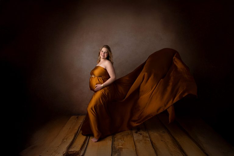 Schwangere Frau im goldenen Kleid mit weit ausgebreiteter Schleppe auf Holzboden, dunkler Hintergrund, strahlendes Lächeln in die Kamera, Babybauchshooting