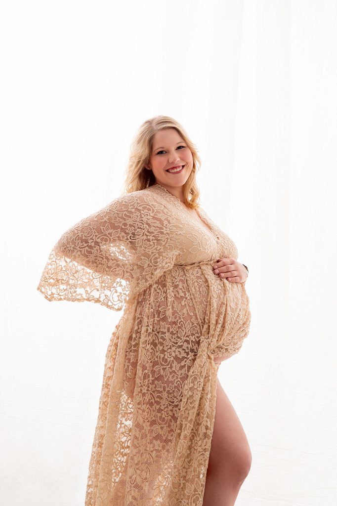 Schwangere Frau steht stolz im Licht vor einem Vorhang, strahlendes Lächeln, Babybauchshooting bei Kahle Fotografie