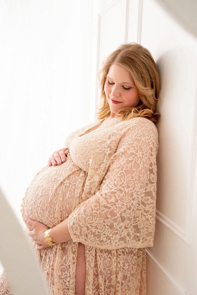 Schwangere Frau lehnt an weißer Kassettenwand, lächelt auf ihren Babybauch, sanftes Licht fällt durch den Vorhang, Babybauchshooting